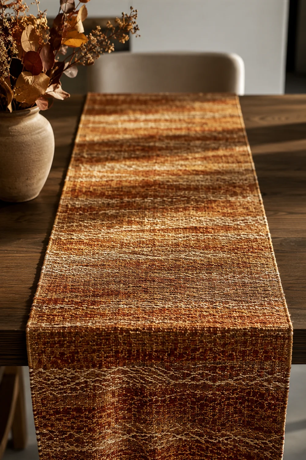 Overhead flat-lay of a rust and gold jacquard-weave table runner on a modern dining table in afternoon light