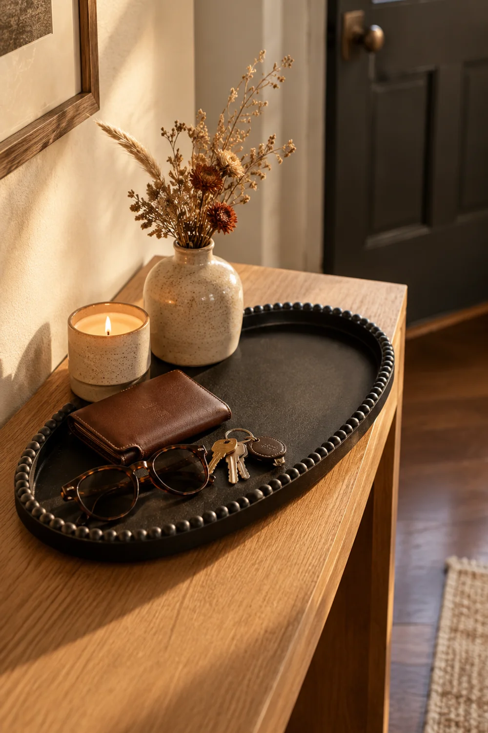 Black matte oval wooden tray with a beaded rim holding keys, sunglasses and a small vase on an entryway console