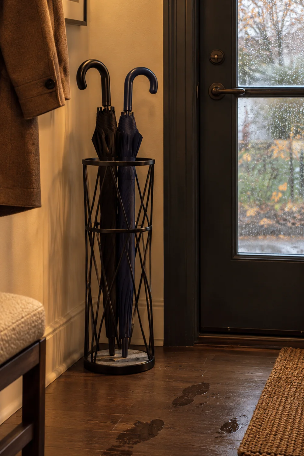 Tall open black metal umbrella holder with a pale stone drip tray holding two umbrellas in the corner of a rainy-day entryway