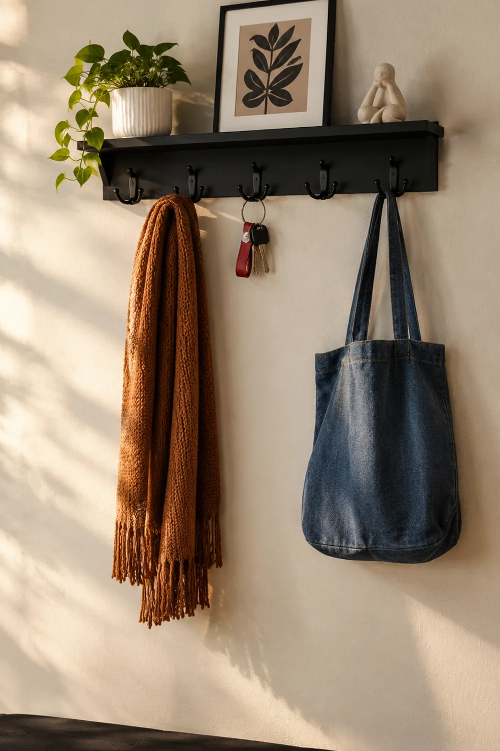Long black wall shelf with five dual hooks holding a scarf and a tote bag with a plant and framed print on the ledge in a small entryway