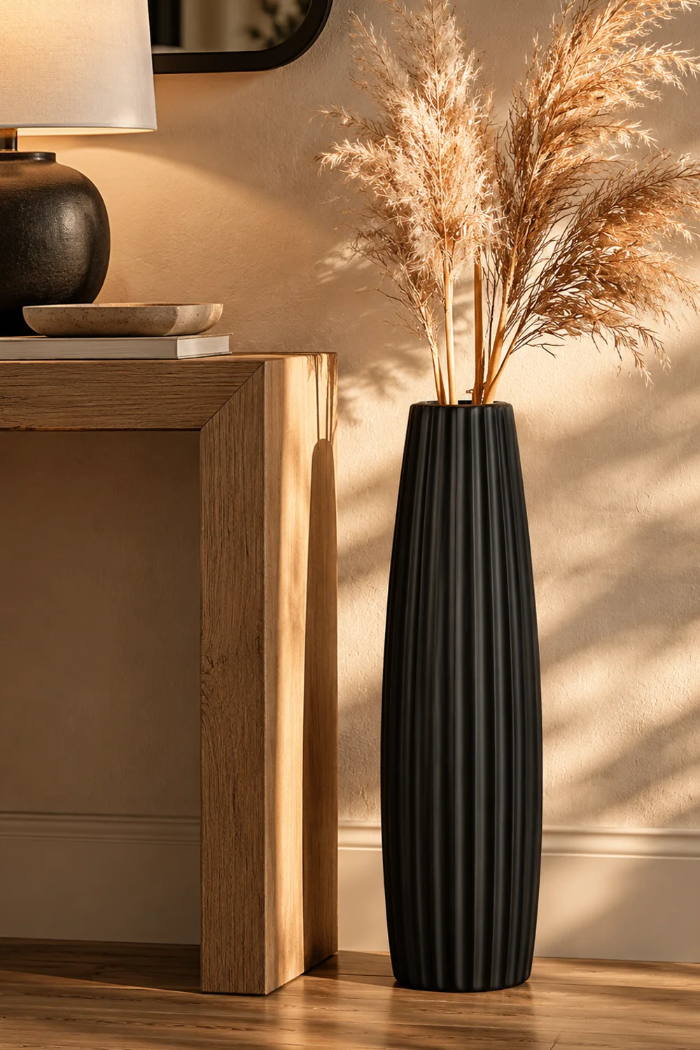 Tall slim matte black ribbed floor vase with dried pampas stems standing beside a console table below the lamp line