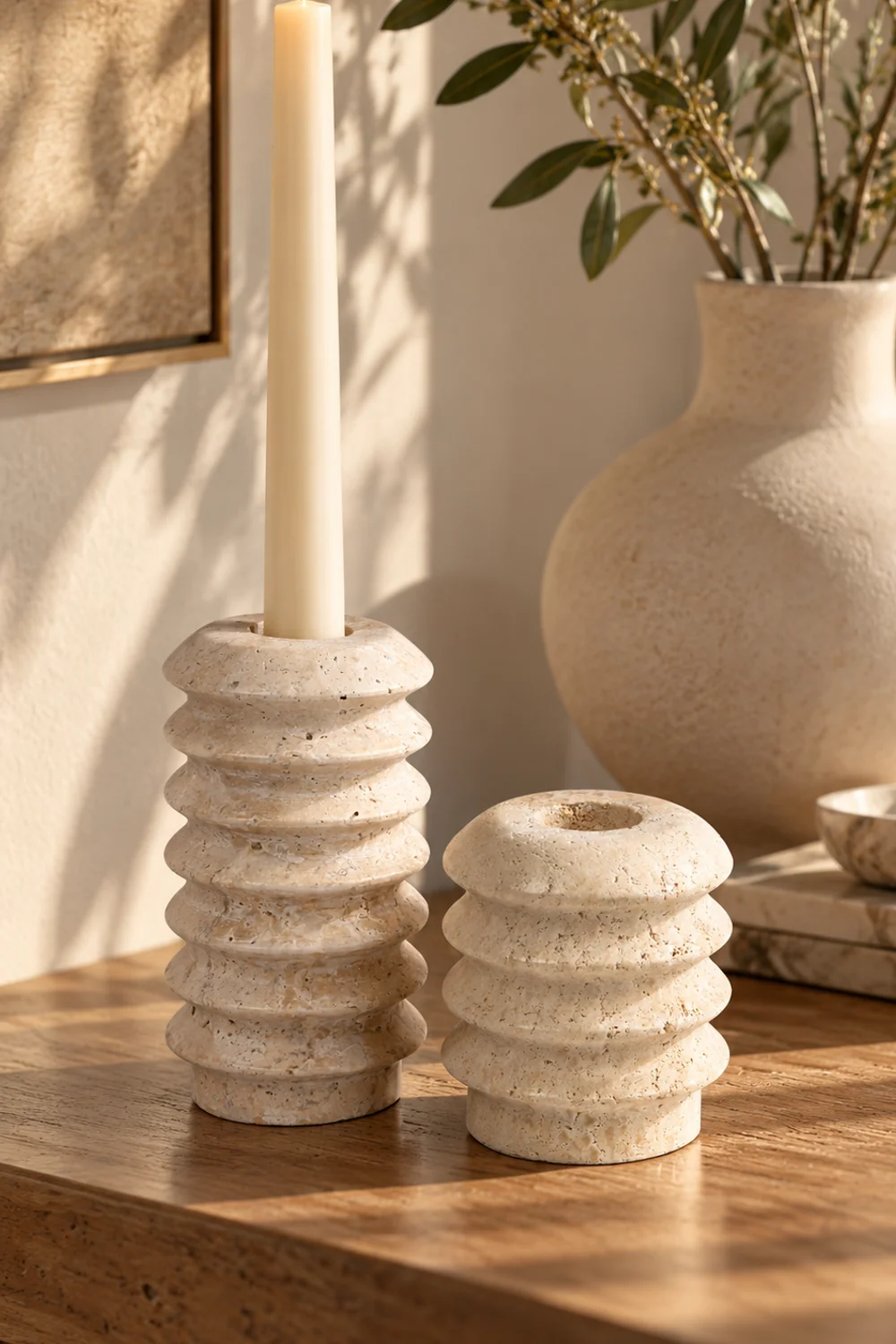 Pair of cream travertine candlesticks in two heights with ivory taper candles on a console table beside a sandstone vase