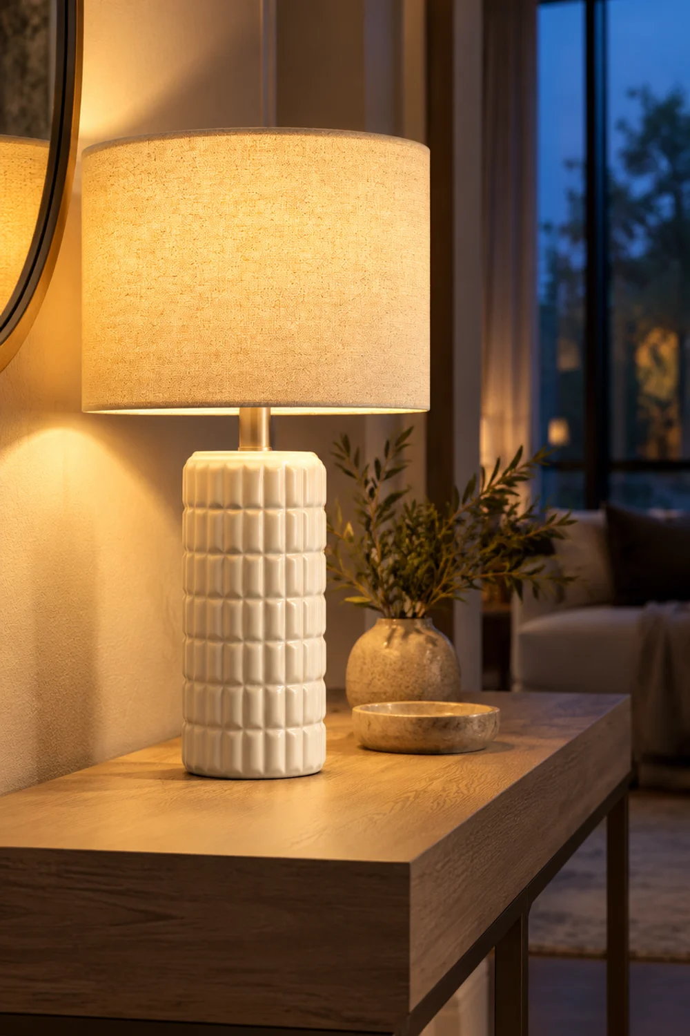 White ceramic table lamp with a grid relief base and a linen drum shade glowing on the left third of a narrow console table at dusk